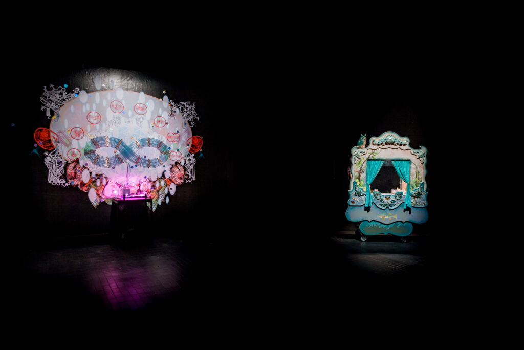 Two art installations in a dark art gallery with black walls. Mounted on the wall on the left is a series of clear tubes in a large figure-eight formation, and underneath them is a wall collage of vinyl decals. On the right is an ornate puppet theatre on wheels, with turquoise curtains and a blue owl perched on top.
