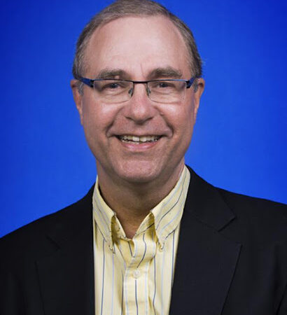 Headshot of man smiling with pale complexion, short grey hair, glasses, yellow button down and dark blazer, against blue backdrop