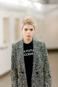 Headshot of a white woman with short spiky blonde hair, wearing a cheetah-print blazer over a black tshirt that says "the future is accessible." The background is blurrred, but appears to be a white room with art on the walls.