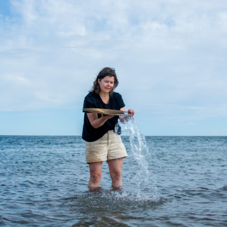 Person with long brown hair, shorts & a tshirt standing knee deep in the ocean, holding a cymbal upside down, balancing water in it like a vessel.