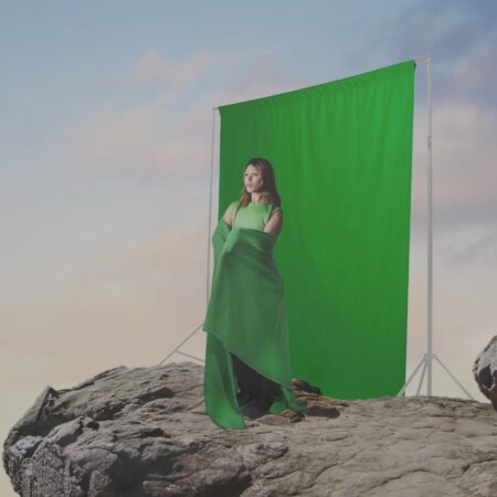 Photoshopped image of a person with long hair & bleached brows, standing on a cliff at dusk. They are dressed in green, with a green blanket wrapped around them, in front of a portable green screen set up on the cliff.