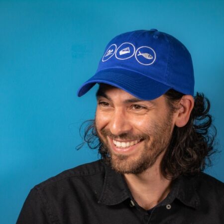 Headshot of young man with dark curly long hair, blue baseball cap against blue backdrop.