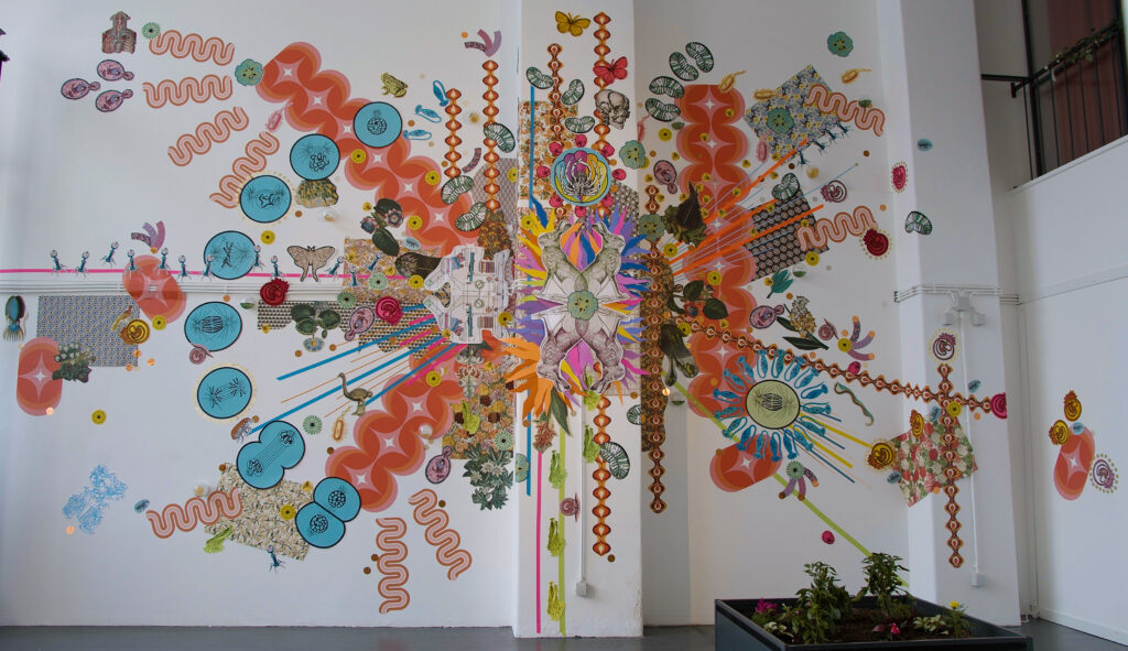 A huge colourful paper collage covers the white wall of an art gallery, and a small temporary garden bed is on the floor with green, yellow, and purple plants. The collage radiates out from the centre, and fetaures bright multicoloured paper cutouts of wallpaper samples, cell diagrams, and illustrations of animals.