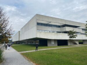 Exterior of a large concrete brutalist building, with blue sky, green lawn, and tree lined sidewalk.