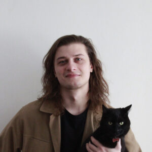 Headshot of person with shoulder-length brown hair staring at camera, wearing brown shirt and holding a black cat wit ha small red bowtie.