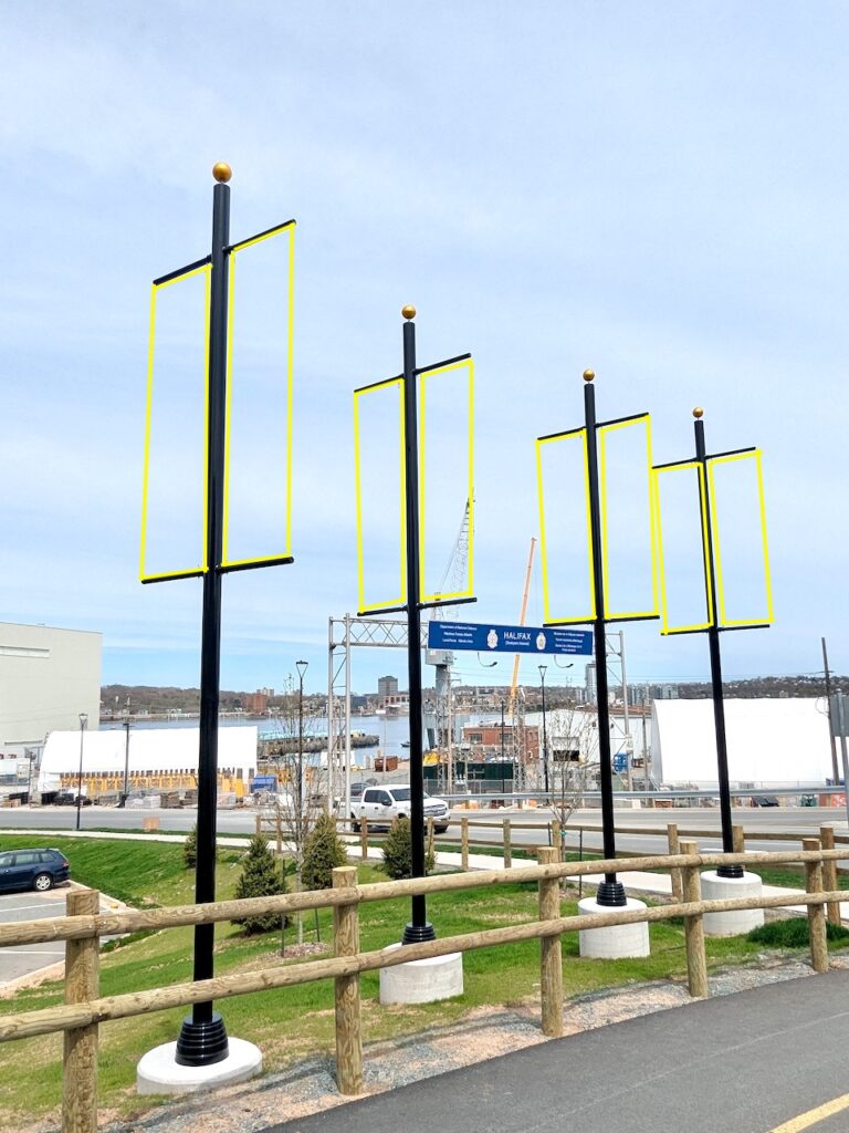 Side of a city street bordered by wood guard rail, and four tall poles, each with a gold ball on top. Four vertical yellow rectangles have been digitally drawn parallel to each lamp post, indicating where a banner would go. In the distance is a highway exit for a navy base, and an ocean harbour.