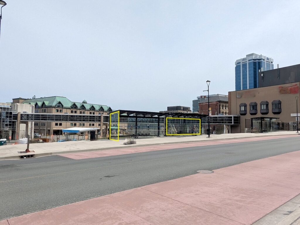 Photo of a cityscape in bright daylight with an overcast sky. Across the street are bus shelters, an overhead pedestrian pedway, and five story buildings. Two yellow rectangles have been digitally drawn over the glass panes of the bus shelters.