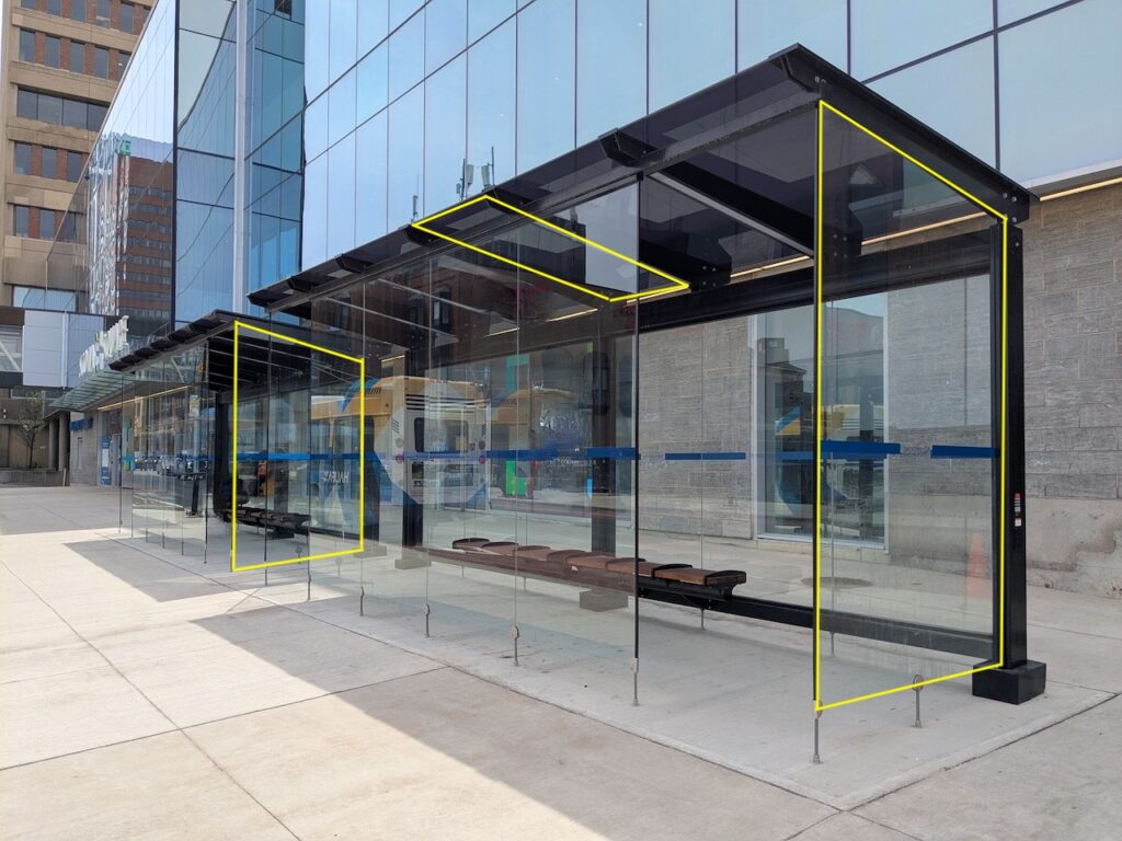 Photo of a glass bus shelter in front of a glass facade building. Three yellow rectangles have been digitally drawn over the glass panes of the bus shelters (two vertical panes on each end, and one pane on the roof of the bus shelter).