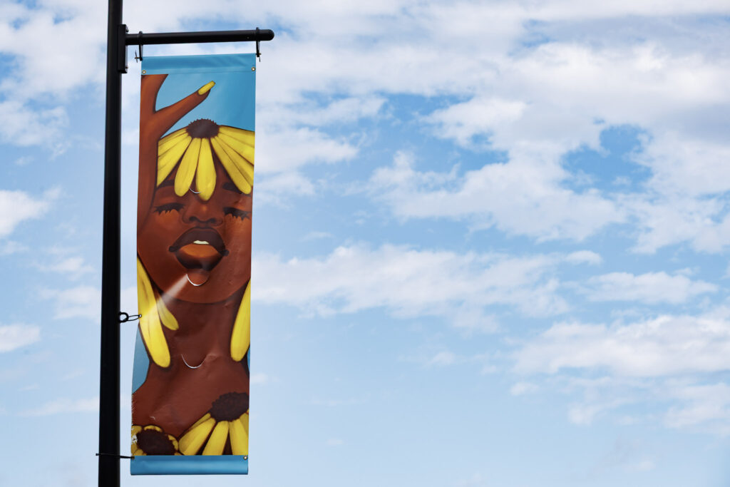 Image of 2025 Interchange artist Carmahn McCalla’s banner “Homegrown” which has a background of a blue sky with clouds. The banner artwork depicts a digital illustration of a Black woman with a deep brown skin tone against a purple background, with eyes closed. She wears a yellow v-neck shirt, gold hoops, coral lipstick, and has white eyebrows. Two white dandelions grow below her, and she has a short white afro that looks like a white dandelion.