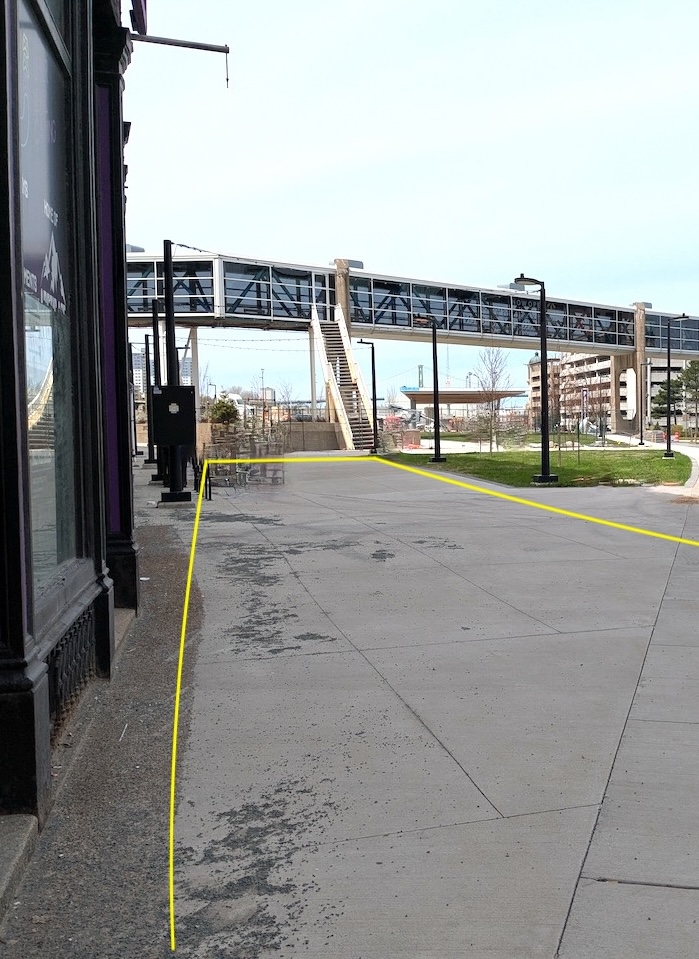Image of the site for the floor vinyl mural, which is a grey cement walk way. The area which is available for design is outlined in a yellow outline, with a loose rectangular shape.