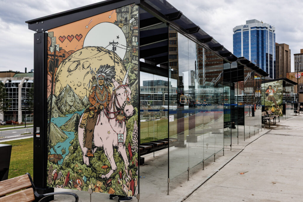 Image is of the transit terminal shelter with artwork from 2025 Interchange artist Zeta Paul, titled “Msɨt No’kmaq (All My Relations): Everything is Smiling”. There is a colourful illustration with an Indigenous man riding a pale pink unicorn in the foreground, wearing a war bonnet, full regalia, and a bow and arrows on his back. He is riding towards the viewer over a hill of green grass covered with mushrooms, teddy bears, lupins, and pink crystals. The unicorn wears a bejewelled harness and around its neck is a medallion with a Mi’kmaw double curve pattern. Over their heads are telephone wires draped across the landscape. The background extends far into the distance where there is a moose at the shoreline of a body of water surrounded by mountains embedded with the faces of Mi’kmaw women wearing peaked caps. On the horizon is a massive full moon rising, and a sun rising above it. Beyond the moon and sun is a high rise apartment building with clotheslines on the fire escapes, there are shooting stars in the sky and in the top left there is three floating red pixellated video game hearts.