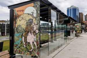 Image is of the transit terminal shelter with artwork from 2025 Interchange artist Zeta Paul, titled “Msɨt No’kmaq (All My Relations): Everything is Smiling”. There is a colourful illustration with an Indigenous man riding a pale pink unicorn in the foreground, wearing a war bonnet, full regalia, and a bow and arrows on his back. He is riding towards the viewer over a hill of green grass covered with mushrooms, teddy bears, lupins, and pink crystals. The unicorn wears a bejewelled harness and around its neck is a medallion with a Mi’kmaw double curve pattern. Over their heads are telephone wires draped across the landscape. The background extends far into the distance where there is a moose at the shoreline of a body of water surrounded by mountains embedded with the faces of Mi’kmaw women wearing peaked caps. On the horizon is a massive full moon rising, and a sun rising above it. Beyond the moon and sun is a high rise apartment building with clotheslines on the fire escapes, there are shooting stars in the sky and in the top left there is three floating red pixellated video game hearts.