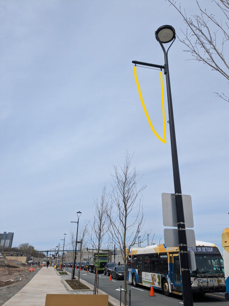 Image of 1 banner pole (with a few in the far background) along Barrington street. These banner poles are empty of banners, in their place are a yellow outline of where the banners will be placed which is in a triangular shape.